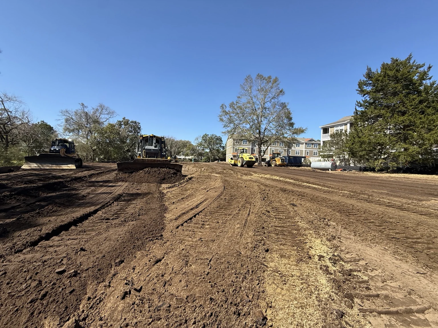 Two bulldozers and a roller on a broad dirt work area beside buildings.