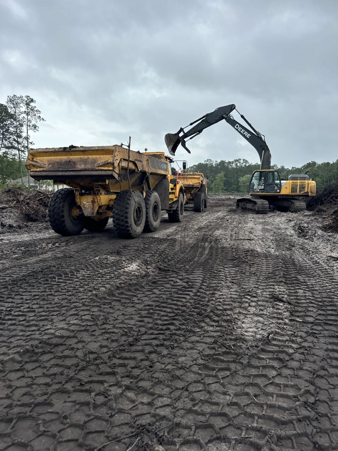 An excavator with its bucket raised above an articulated dump truck on an earthwork site, with another truck behind.