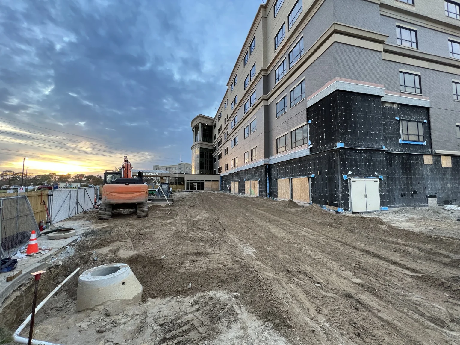 An excavator and exposed utility access structures in an earthwork area beside a multistory building at sunset.